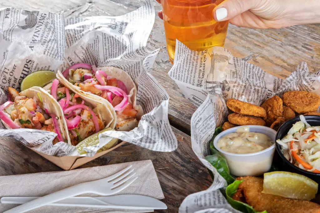 A rustic table setting with a person holding a glass of amber-colored drink, next to food items including fish tacos with pickled onions and a basket of fried food accompanied by dipping sauces and coleslaw.